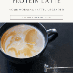 Creamy protein latte with swirled foam in a black ceramic mug, styled on a dark countertop with a handheld milk frother and a protein powder scoop nearby.