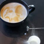 Creamy protein latte with swirled foam in a black ceramic mug, styled on a dark countertop with a handheld milk frother and a protein powder scoop nearby.