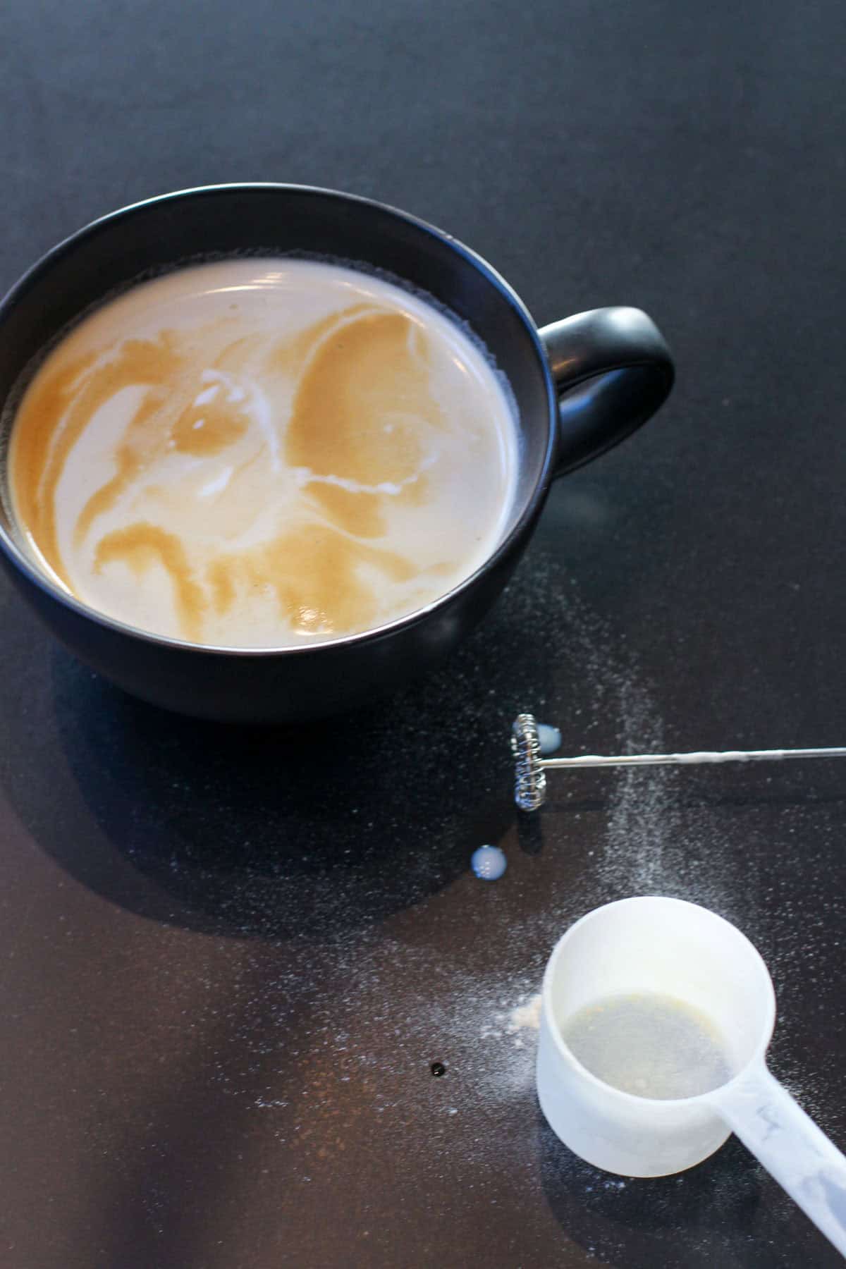 Creamy protein latte with swirled foam in a black ceramic mug, styled on a dark countertop with a handheld milk frother and a protein powder scoop nearby.