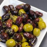 Warm marinated olives made with Kalamata, green, and black olives tossed in olive oil, lemon zest, and herbs de Provence, served in a white dish.