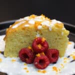Slice of orange olive oil cake topped with whipped honey ricotta, fresh raspberries, and citrus zest on a dark plate