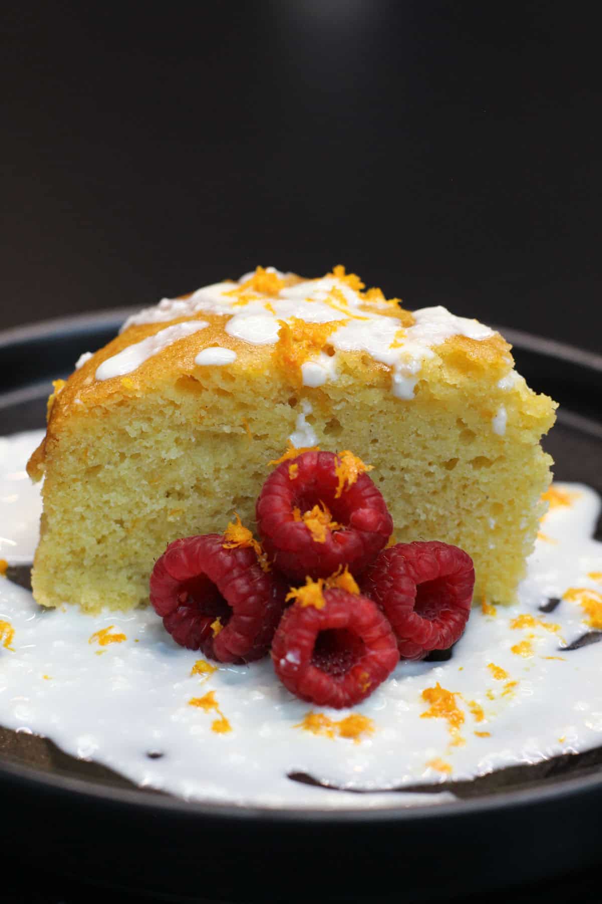 Slice of orange olive oil cake topped with whipped honey ricotta, fresh raspberries, and citrus zest on a dark plate