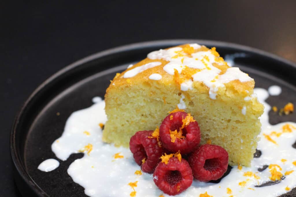 Slice of orange olive oil cake topped with whipped honey ricotta, fresh raspberries, and citrus zest on a dark plate