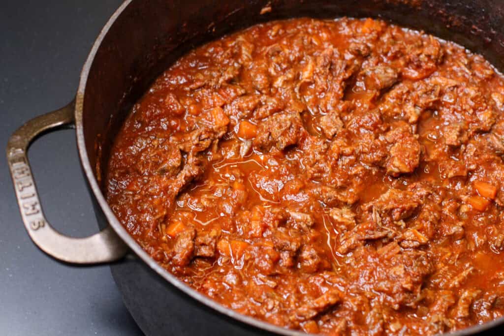 Slow-braised beef ragú simmering in a cast iron Dutch oven, with tender shredded beef in a rich tomato and red wine sauce.