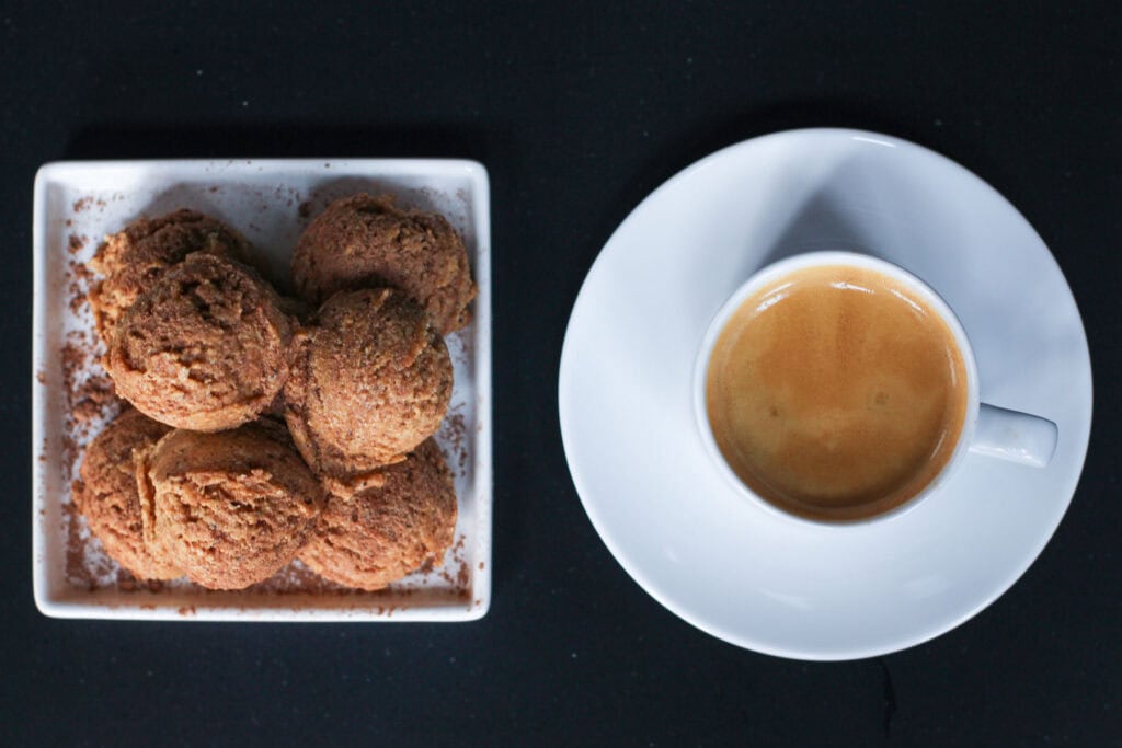 Tiramisu protein bites stacked on a white ceramic dish, lightly dusted with cocoa powder, showing a soft, espresso-speckled texture against a dark background. There is also a white cup and saucer of espresso next to the bites.