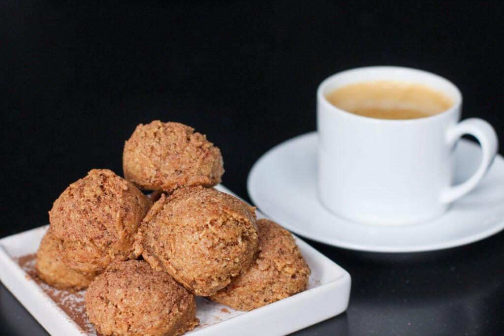 Tiramisu protein bites stacked on a white ceramic dish, lightly dusted with cocoa powder, showing a soft, espresso-speckled texture against a dark background. There is also a white cup and saucer of espresso next to the bites.