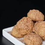 Tiramisu protein bites stacked on a white ceramic dish, lightly dusted with cocoa powder, showing a soft, espresso-speckled texture against a dark background. There is also a white cup and saucer of espresso next to the bites.