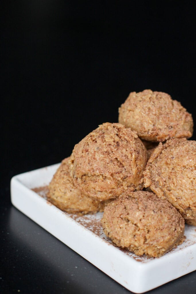 Tiramisu protein bites stacked on a white ceramic dish, lightly dusted with cocoa powder, showing a soft, espresso-speckled texture against a dark background. There is also a white cup and saucer of espresso next to the bites.