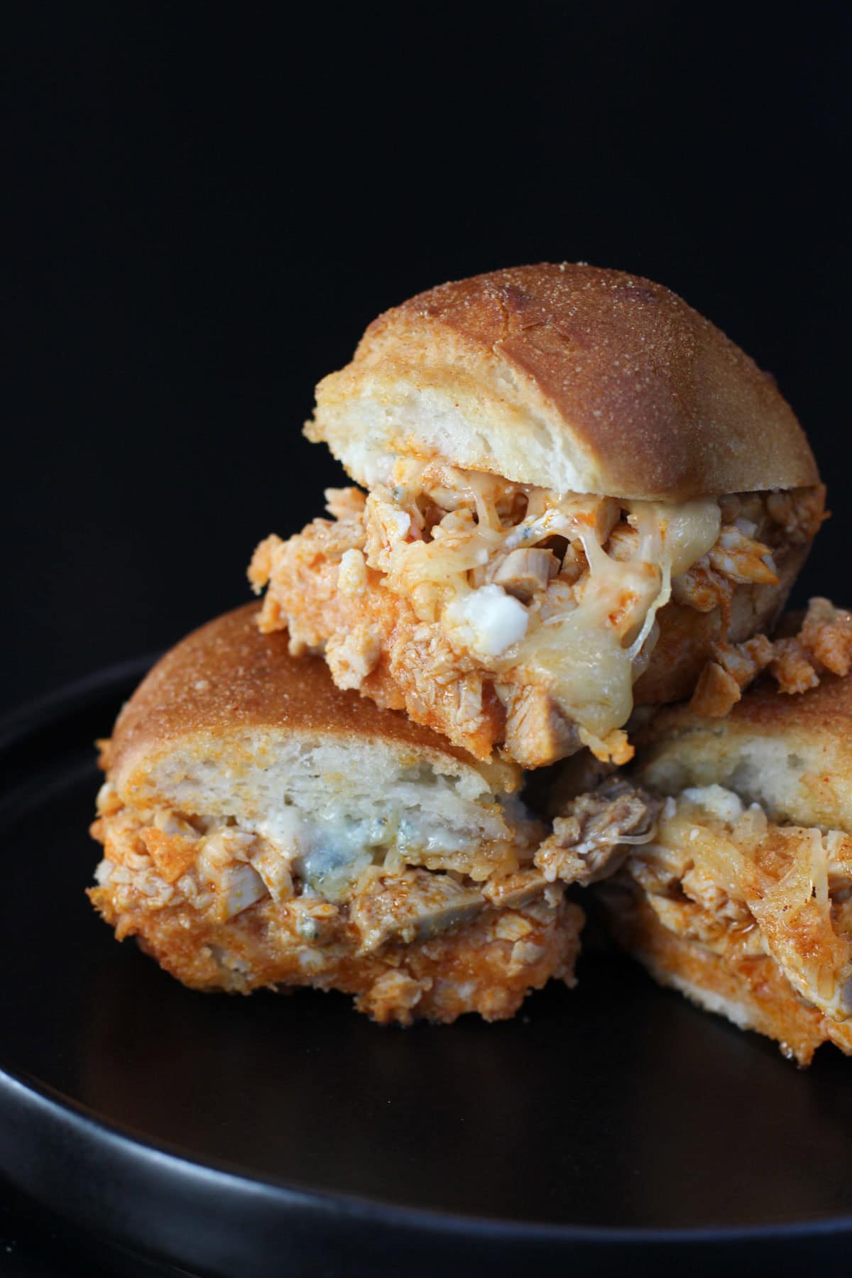 Baked buffalo chicken sliders on Hawaiian rolls with melted cheese and garlic butter tops stacked on black plate.