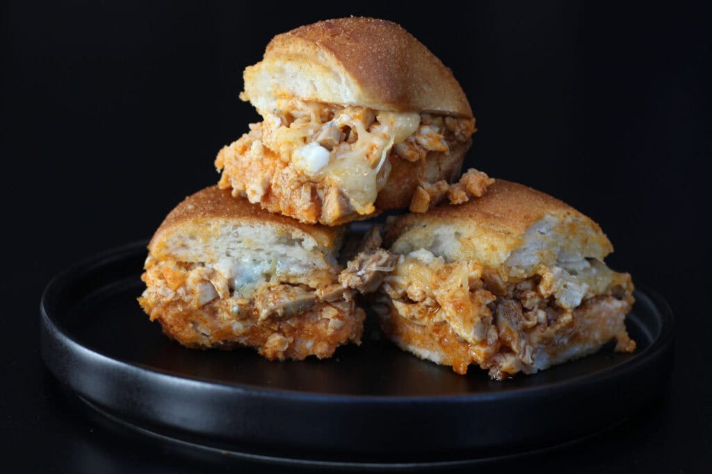 Baked buffalo chicken sliders on Hawaiian rolls with melted cheese and garlic butter tops stacked on black plate.