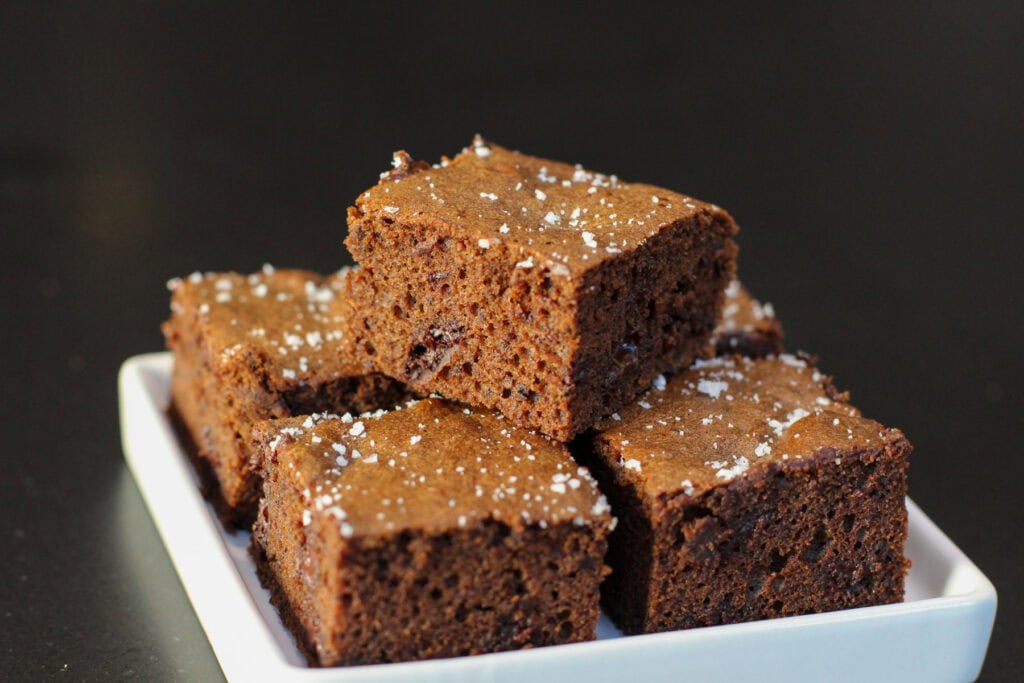Fudgy chocolate sea salt brownies topped with flaky sea salt, sliced into squares on white serving dish.