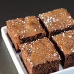 Fudgy chocolate sea salt brownies topped with flaky sea salt, sliced into squares on white serving dish.