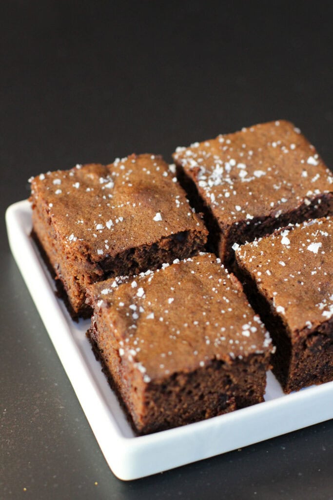 Fudgy chocolate sea salt brownies topped with flaky sea salt, sliced into squares on white serving dish.