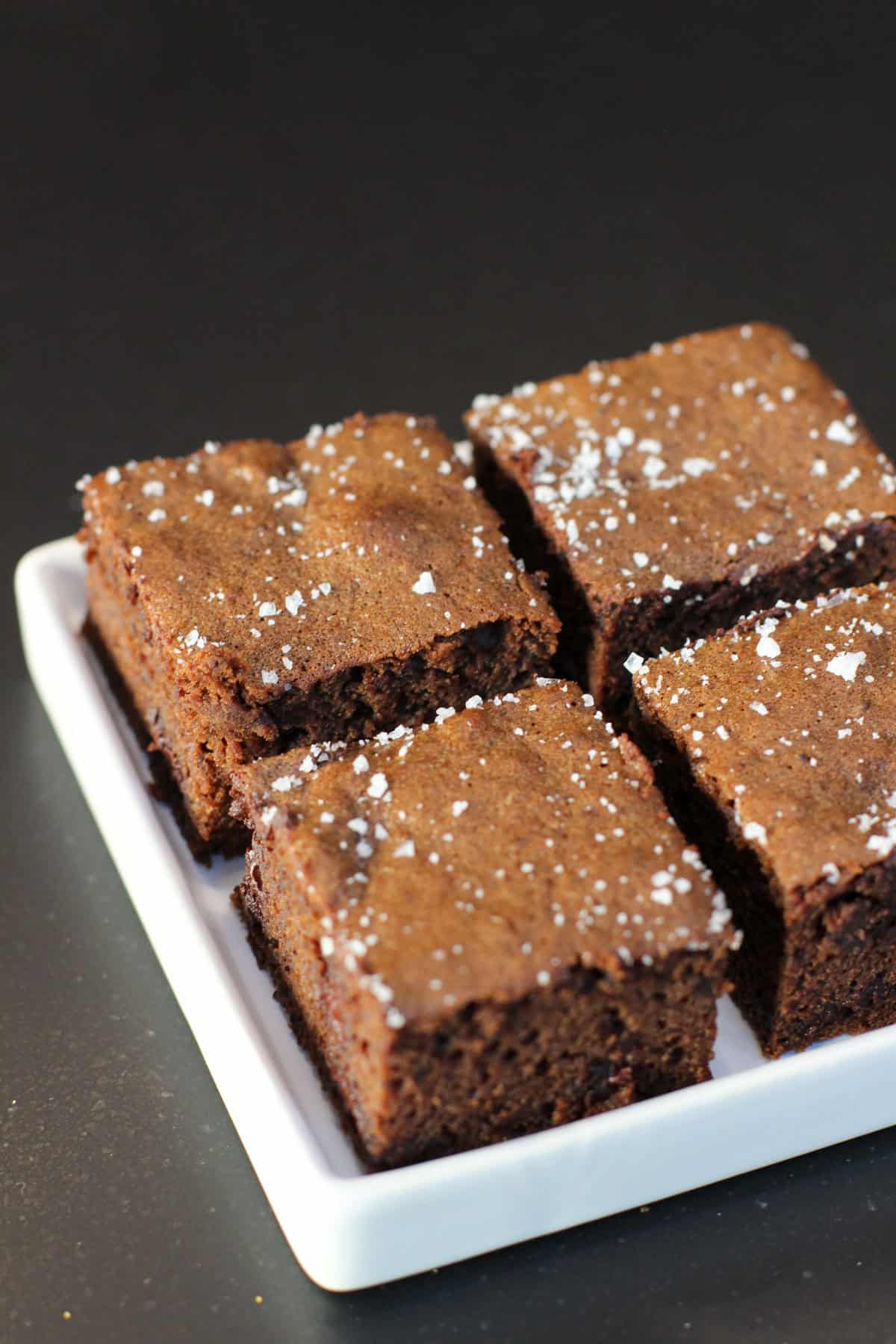 Fudgy chocolate sea salt brownies topped with flaky sea salt, sliced into squares on white serving dish.