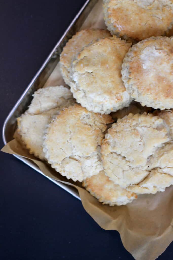 Golden gluten-free biscuits stacked in a metal baking pan lined with parchment paper, showing flaky edges and a soft, rustic texture.