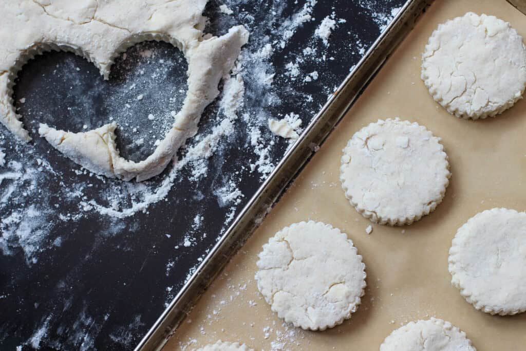 Gluten-free biscuit dough rolled out and cut into rounds on a floured surface, with unbaked biscuits arranged on a parchment-lined baking sheet.