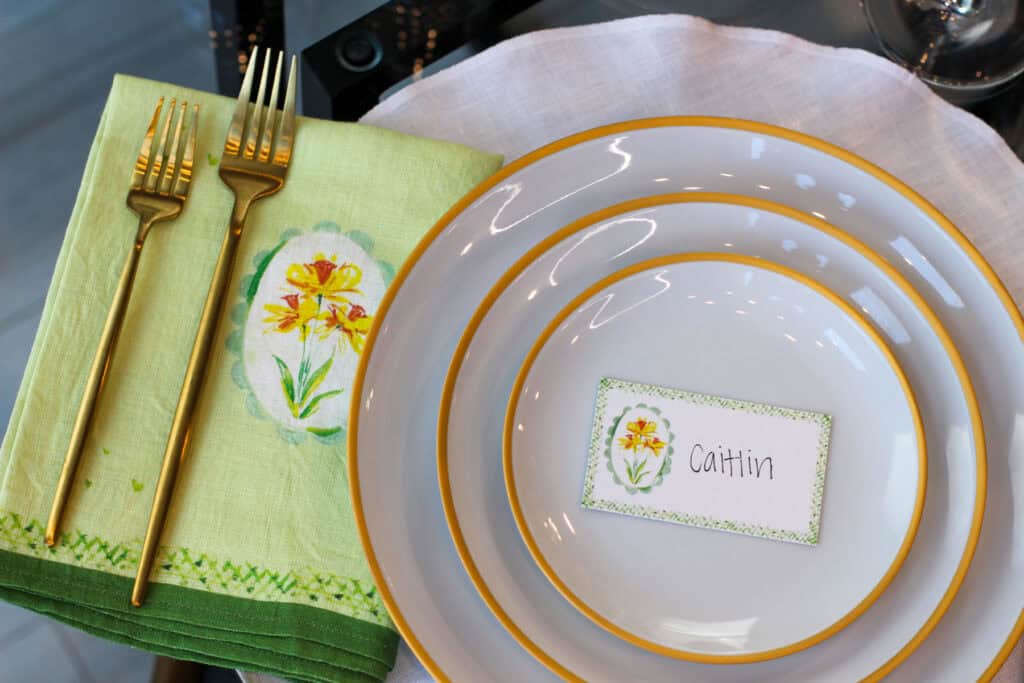 Elegant spring place setting with layered plates, green linen napkin, gold flatware, and personalized place card