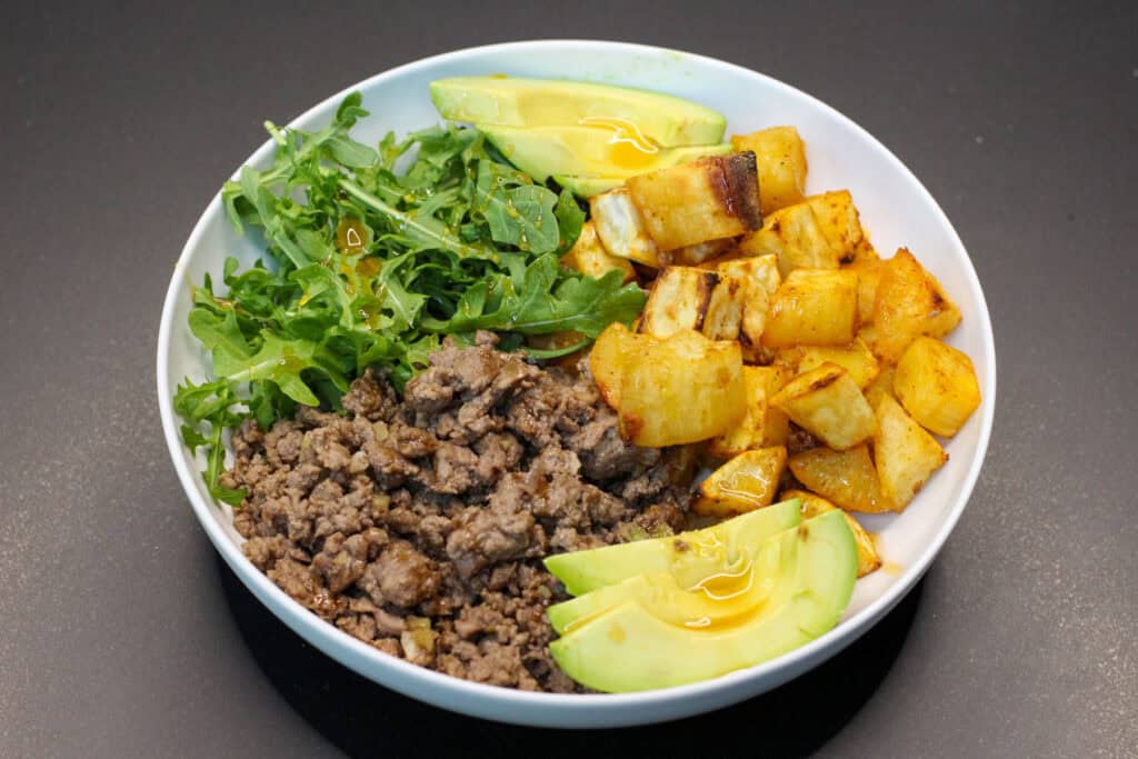 Hot honey ground venison sweet potato bowl with avocado, arugula, and roasted sweet potatoes in a white bowl
