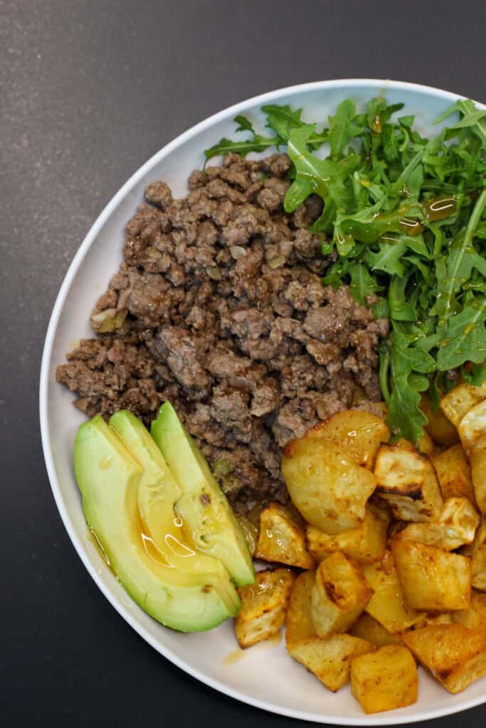 Hot honey ground venison sweet potato bowl with avocado, arugula, and roasted sweet potatoes in a white bowl