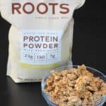 Crunchy homemade protein granola served in a glass bowl with Redmond Roots vanilla protein powder in the background