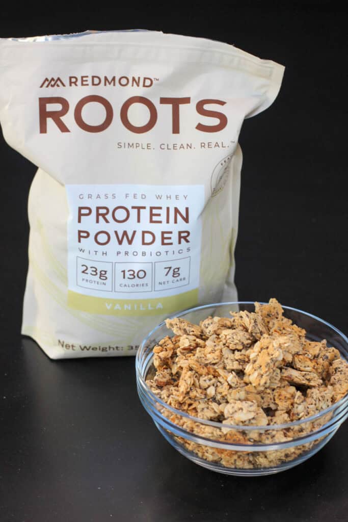 Crunchy homemade protein granola served in a glass bowl with Redmond Roots vanilla protein powder in the background