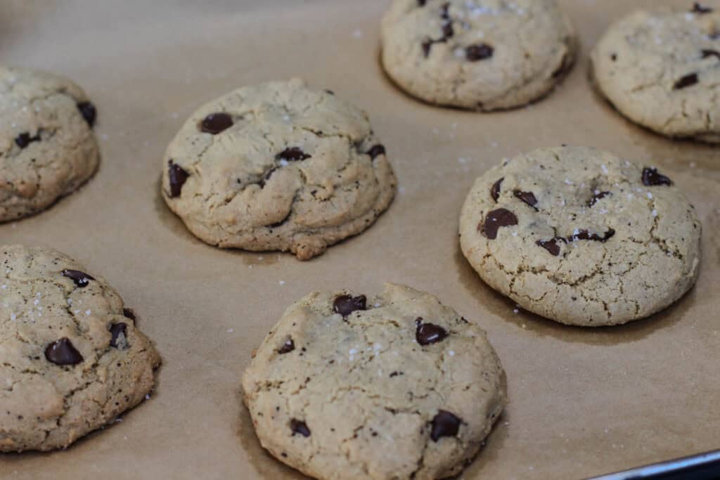 Freshly baked sea salt espresso chocolate chip cookies cooling on a parchment-lined baking sheet with chocolate chips and flaky sea salt.