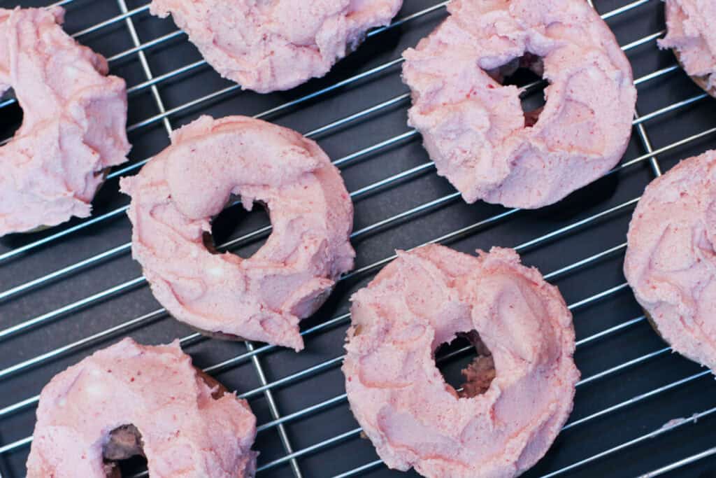 Freshly frosted strawberry buttercream donuts cooling on a wire baking rack.