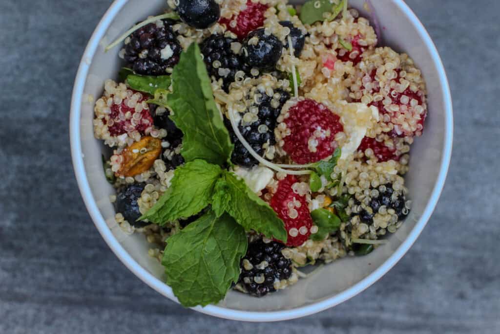 Summer Berry Quinoa Salad with fresh raspberries, blueberries, blackberries, feta cheese, pistachios, mint, and citrus honey dressing served in a white bowl.